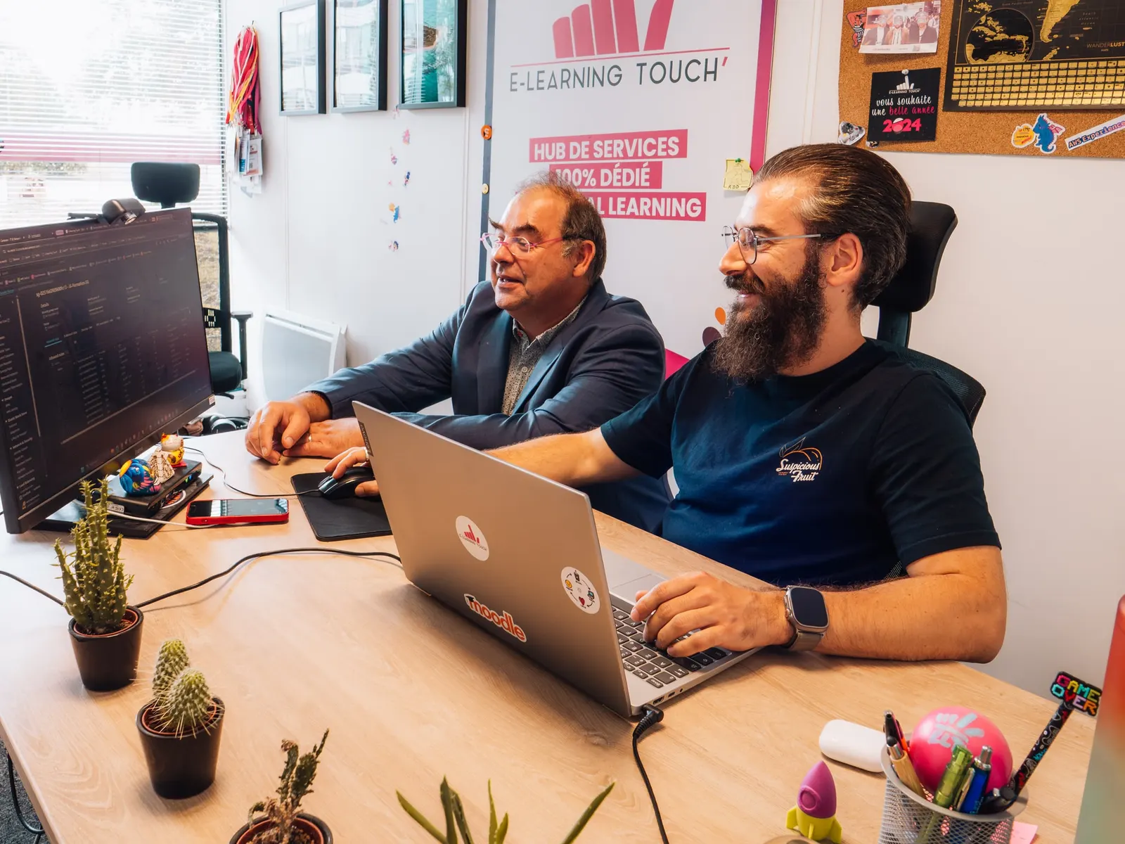 Deux collègues d'E-learning Touch' travaillent côte à côte sur un ordinateur portable, dans les bureaux décorés aux couleurs de l'entreprise.
