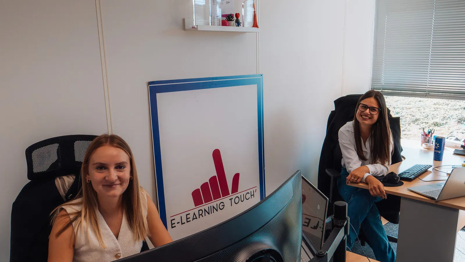 Deux collègues d'E-learning Touch' sourient depuis leur poste de travail, devant le logo de l'entreprise.