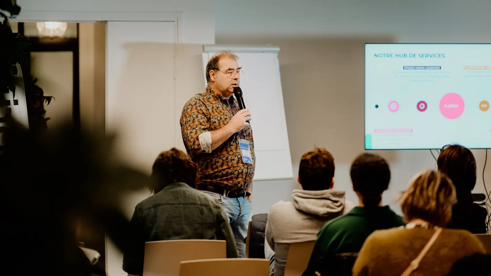 Un intervenant d'E-learning Touch' présente au micro devant un public assis, une diapositive projetée à côté de lui.